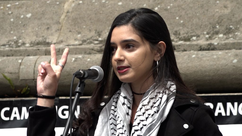 A young female adult with armpit length black hair and wearing a Palestinian keffiyeh (a chequered black and white scarf) holds up the peace sign whilst standing at a microphone. 