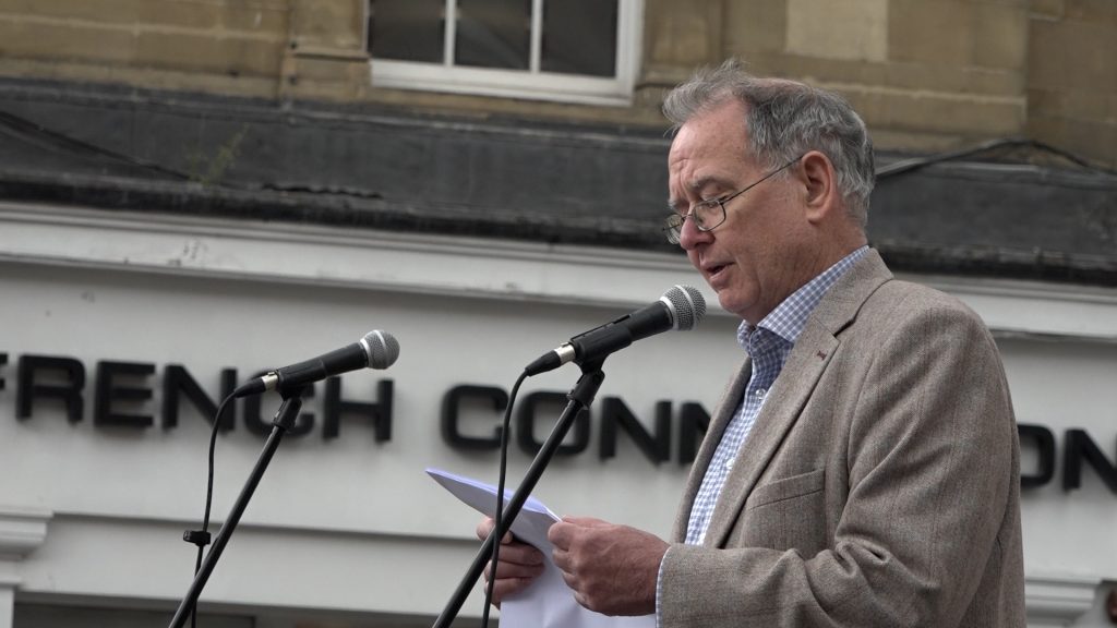An older male adult with short grey hair and glasses wearing a grey blazer and light shirt stands at a microphone and reads from sheets of papers. 