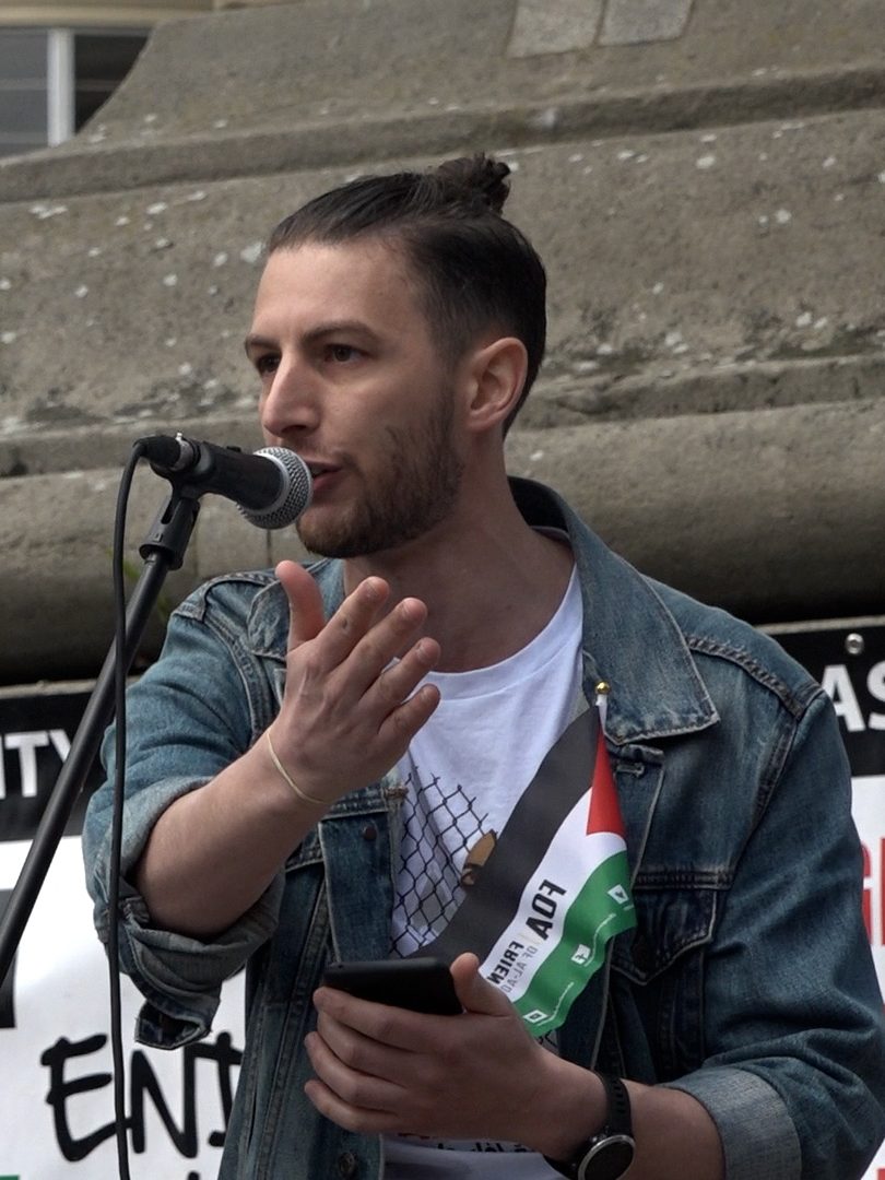 A young male adult with brown hair in a small bun and light facial hair stands at a microphone gesturing out to the crowd. He has a small Palestinian flag in his denim top breast pocket which has the logo of the Friends of Al-Awsa printed on it. 